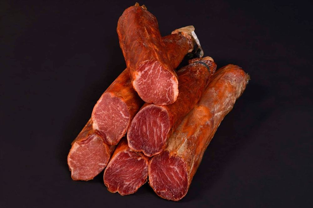 Rebanadas de lomo de cerdo curado, de color marrón rojizo con veteado visible, apiladas sobre una superficie negra.