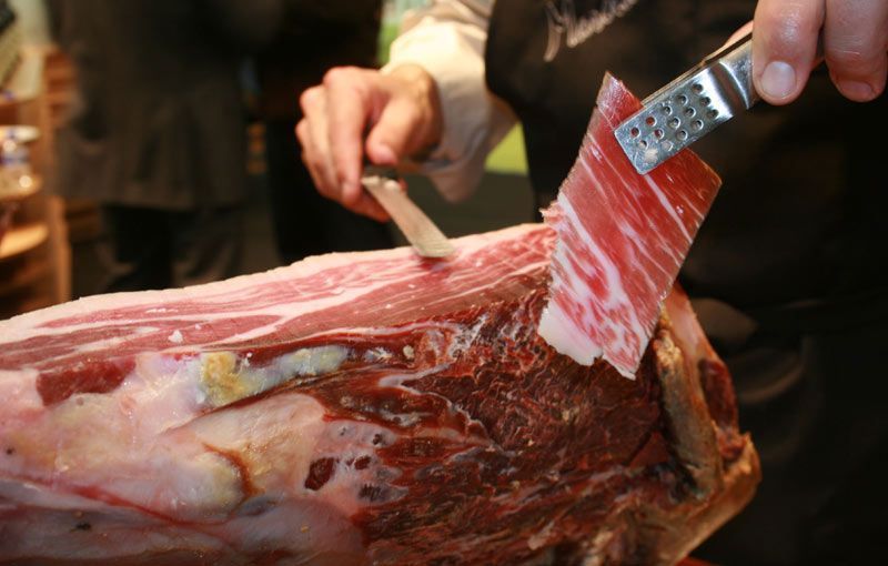 Jamón curado parcialmente loncheado, montado sobre un soporte con tabla de corte de madera.