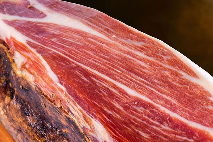 Primer plano de un jamón curado que muestra carne roja con vetas blancas, apoyada sobre una superficie más oscura.