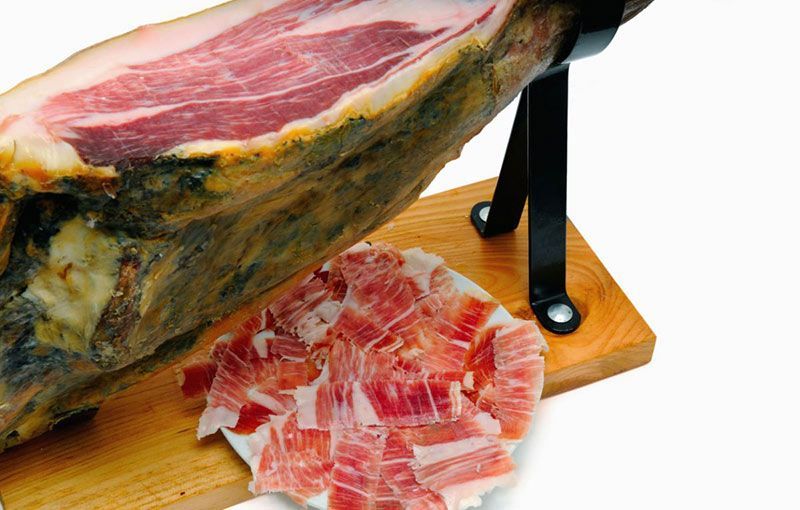 Jamón curado parcialmente loncheado, montado sobre un soporte con tabla de corte de madera.