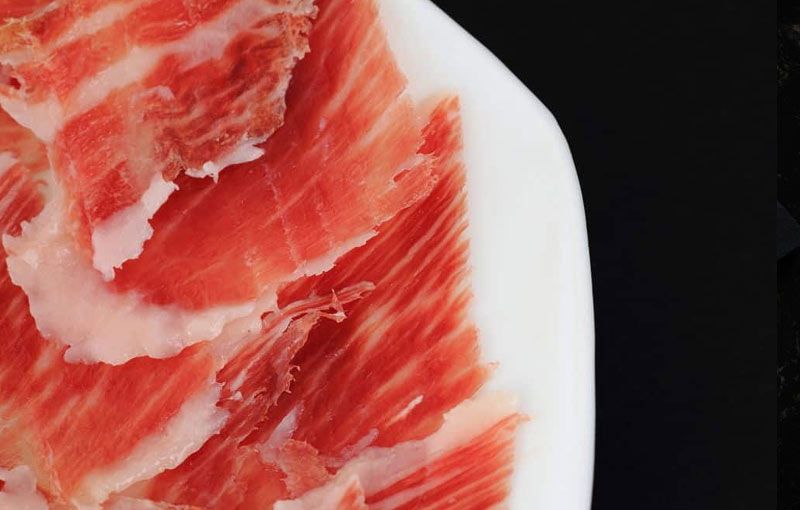 Jamón ibérico cortado en lonchas finas con grasa roja y blanca jaspeada sobre un plato blanco sobre un fondo negro.