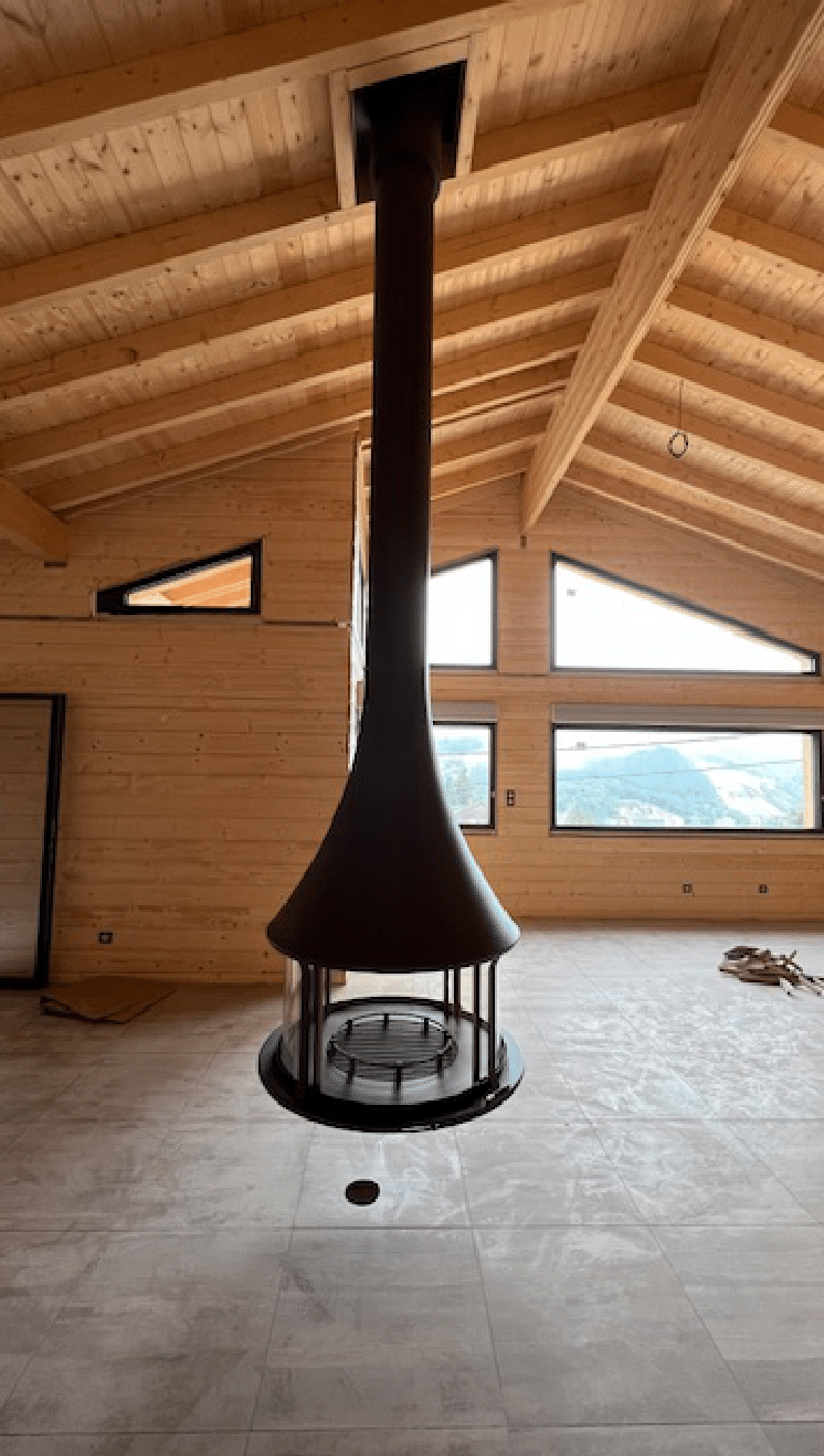 Intérieur de chalet moderne avec une cheminée noire suspendue et de grandes fenêtres offrant une vue sur la montagne