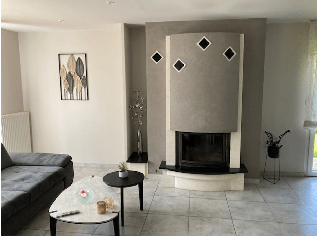 Salon moderne avec un canapé gris, une table basse et une cheminée centrale sur un sol carrelé.