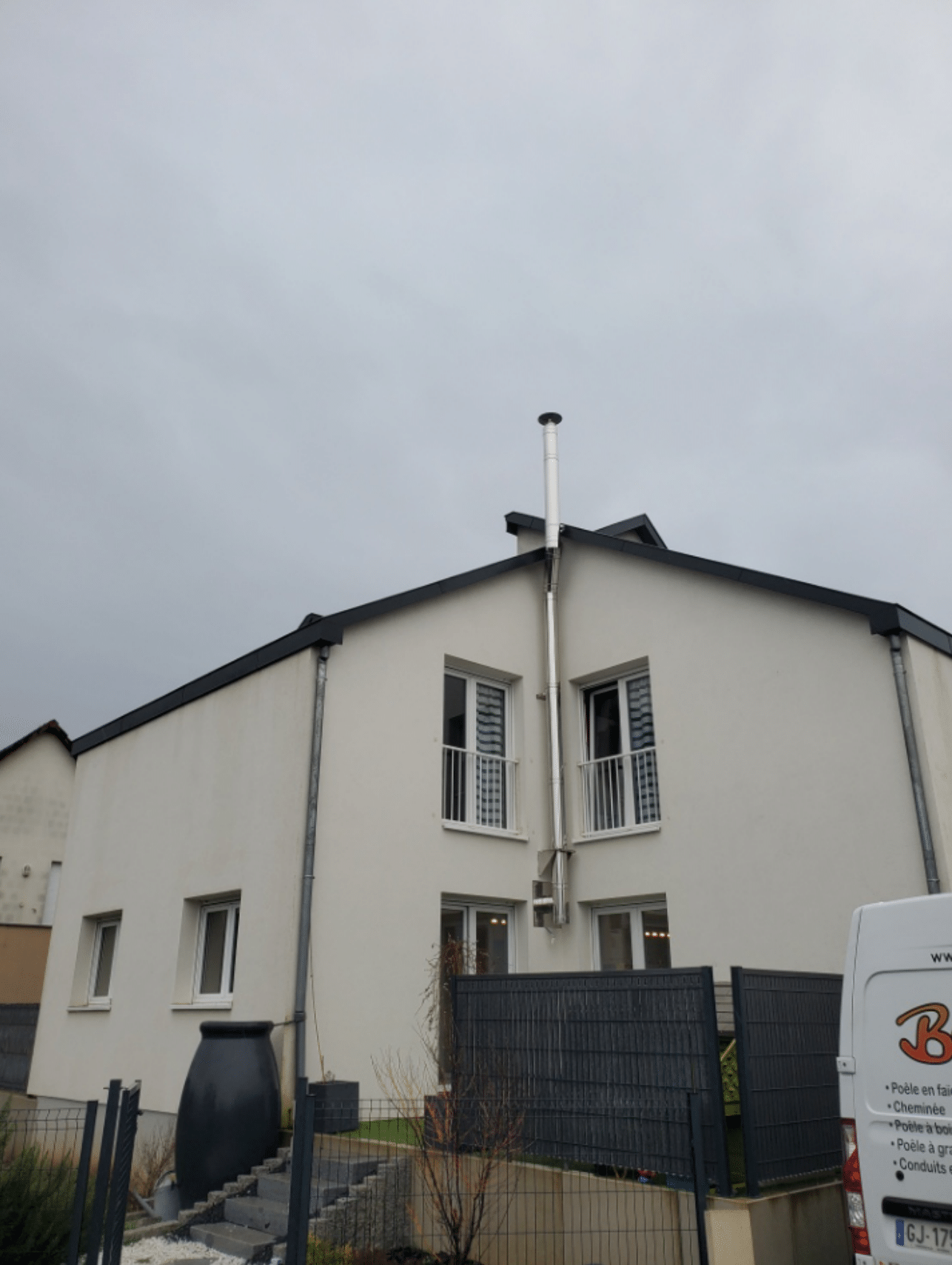 Maison blanche à deux étages avec toit noir, fenêtres à barreaux et cour avant clôturée sous un ciel nuageux