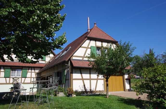 Maison blanche traditionnelle avec toit de tuiles rouges, volets verts et jardin sous un ciel bleu ensoleillé