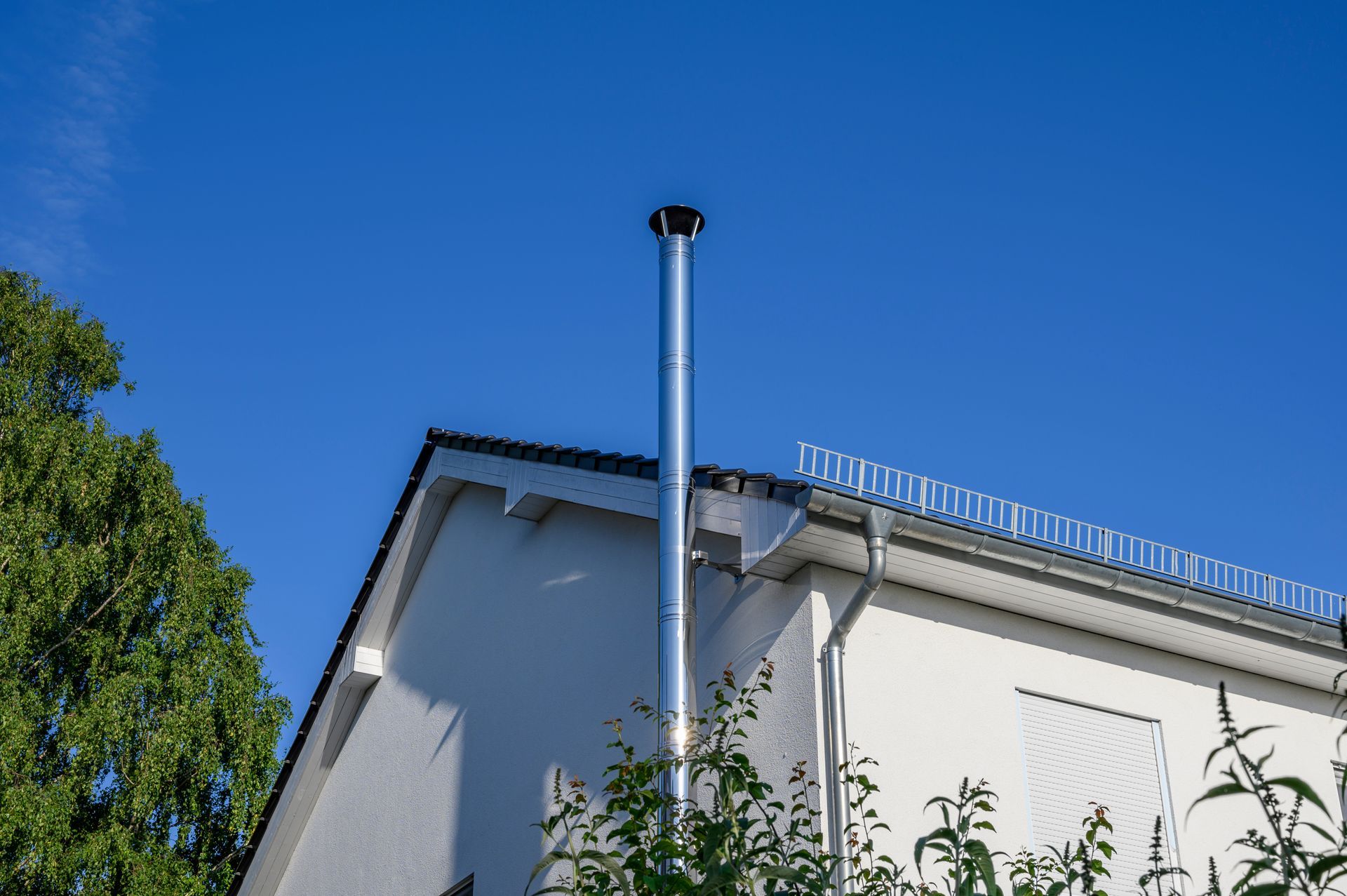 Toit d'un immeuble blanc avec une haute cheminée se détachant sur un ciel bleu clair, encadré par des arbres verdoyants