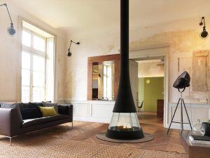 Salon minimaliste avec de grandes fenêtres, un canapé noir, du parquet et une cheminée suspendue centrale.