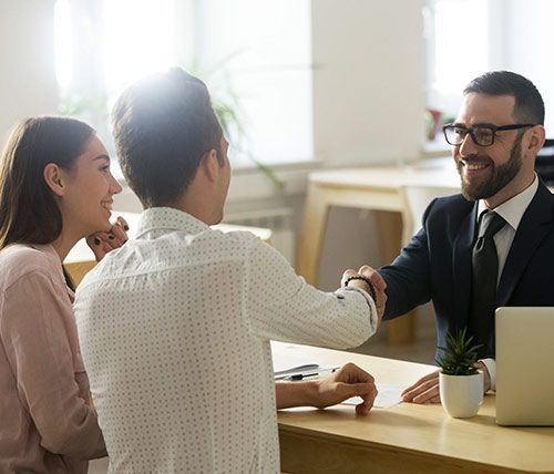 Un profesional vestido de traje estrecha la mano de un cliente al otro lado de un escritorio, mientras otra persona observa; todos sonríen en una oficina.
