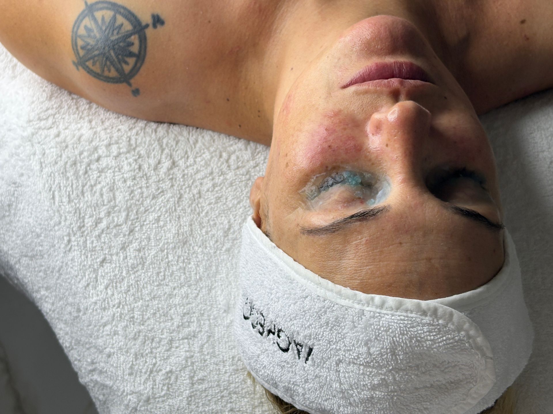Persona con los ojos cerrados y una diadema de spa. Maquillaje verde azulado visible y tatuaje en el hombro. Tumbada sobre una toalla blanca.