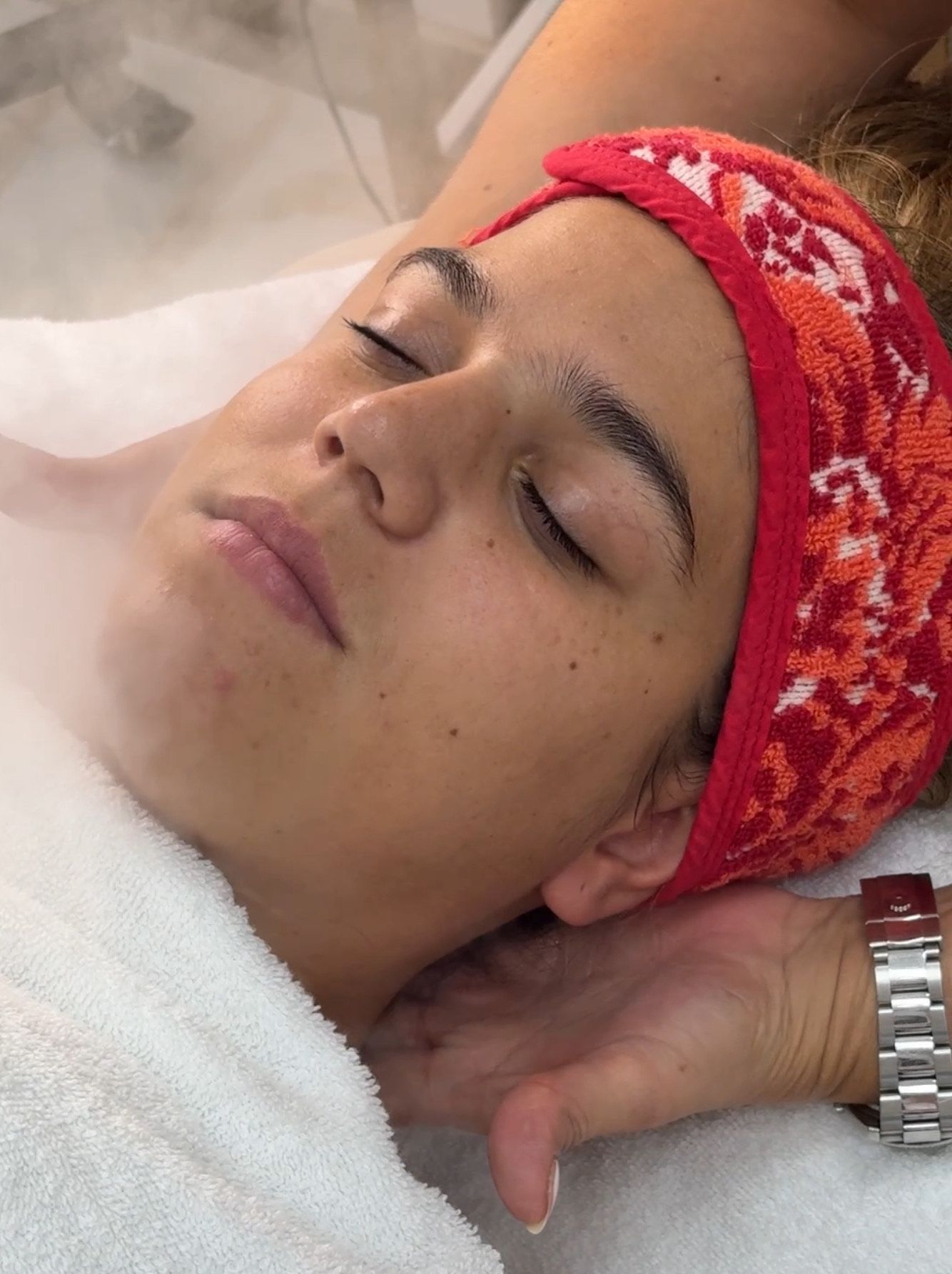Mujer recibiendo tratamiento de vapor facial, con los ojos cerrados y luciendo una diadema roja.