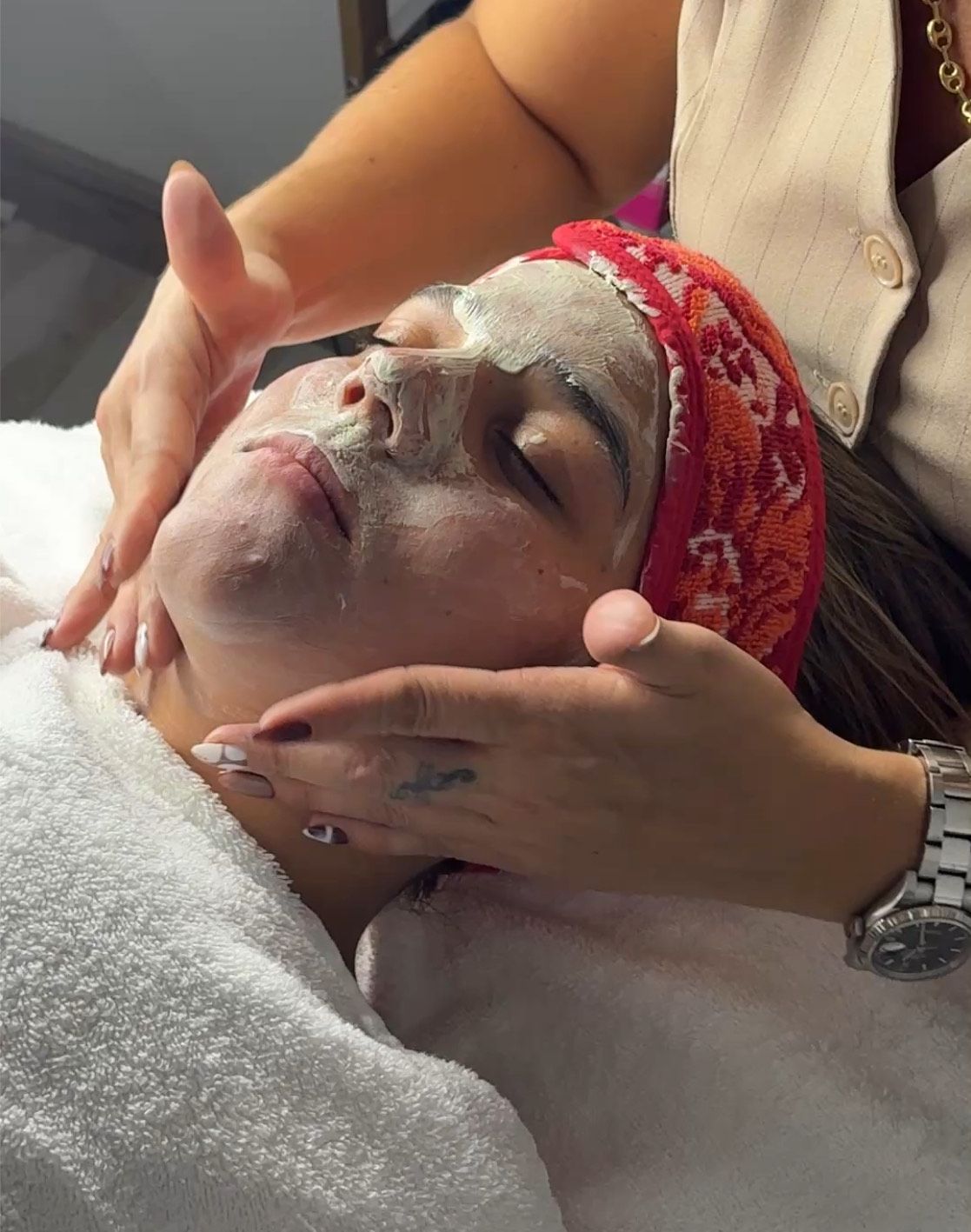 Mujer recibiendo tratamiento facial, con las manos masajeando suavemente el rostro, mascarilla aplicada, con diadema, en un ambiente interior.
