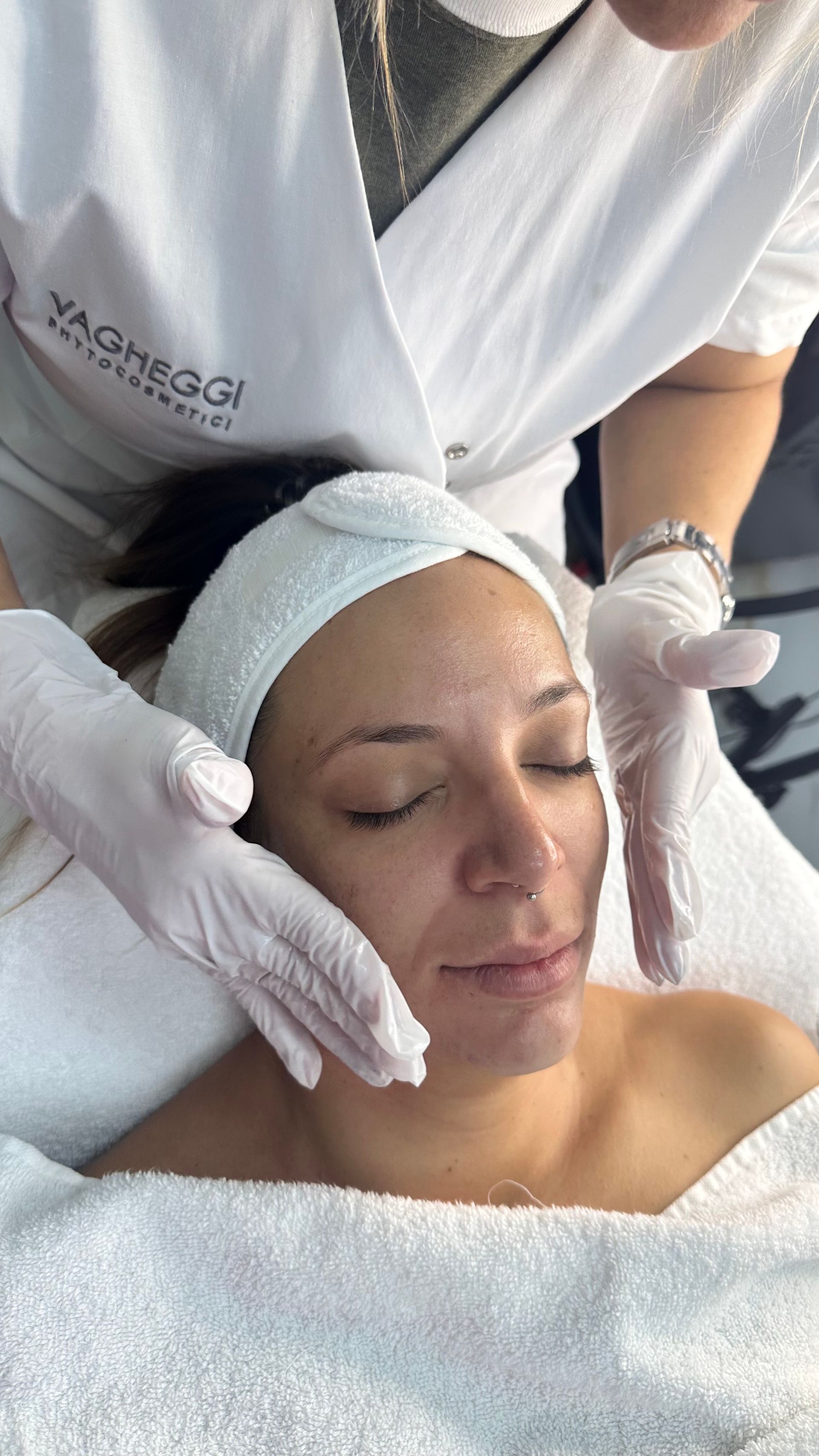 Mujer recibiendo tratamiento facial, ojos cerrados, manos en la cara, usando diadema y guantes.
