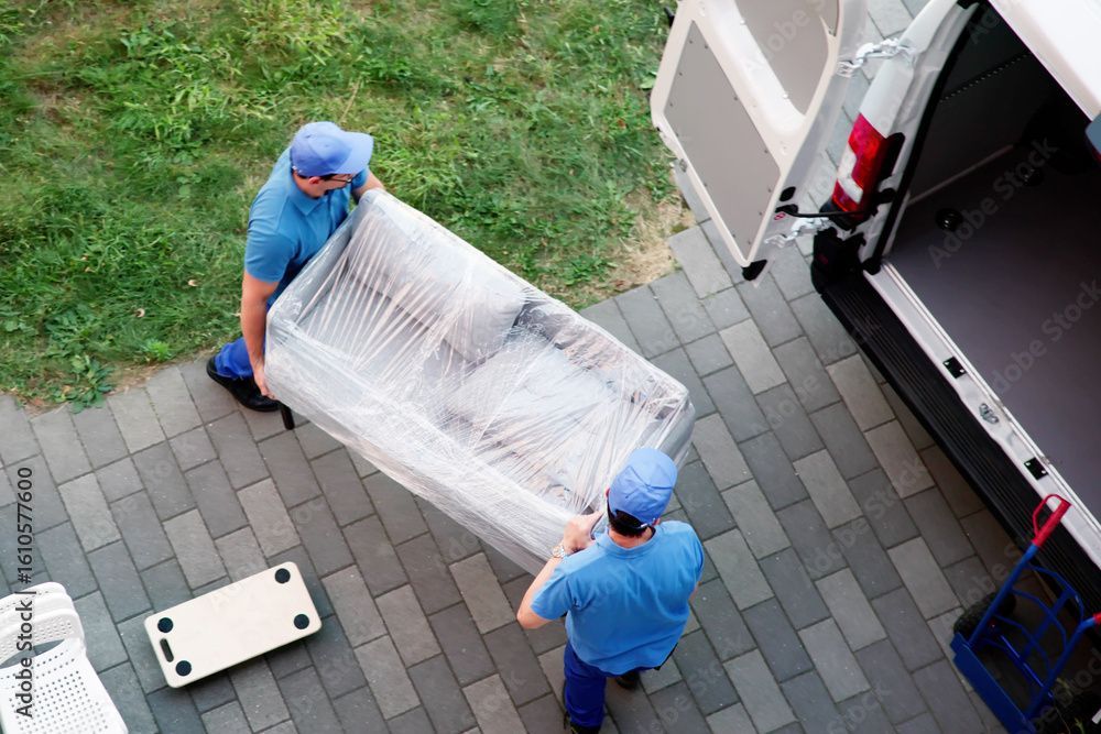 Zwei Umzugshelfer in blauen Uniformen laden auf einer gemauerten Terrasse eine verpackte Couch aus einem weissen Lieferwagen ab.