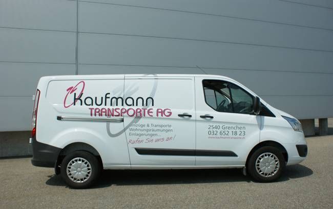 Ein weisser Lieferwagen der Kaufmann Transporte AG parkt vor einem grauen Gebäude.