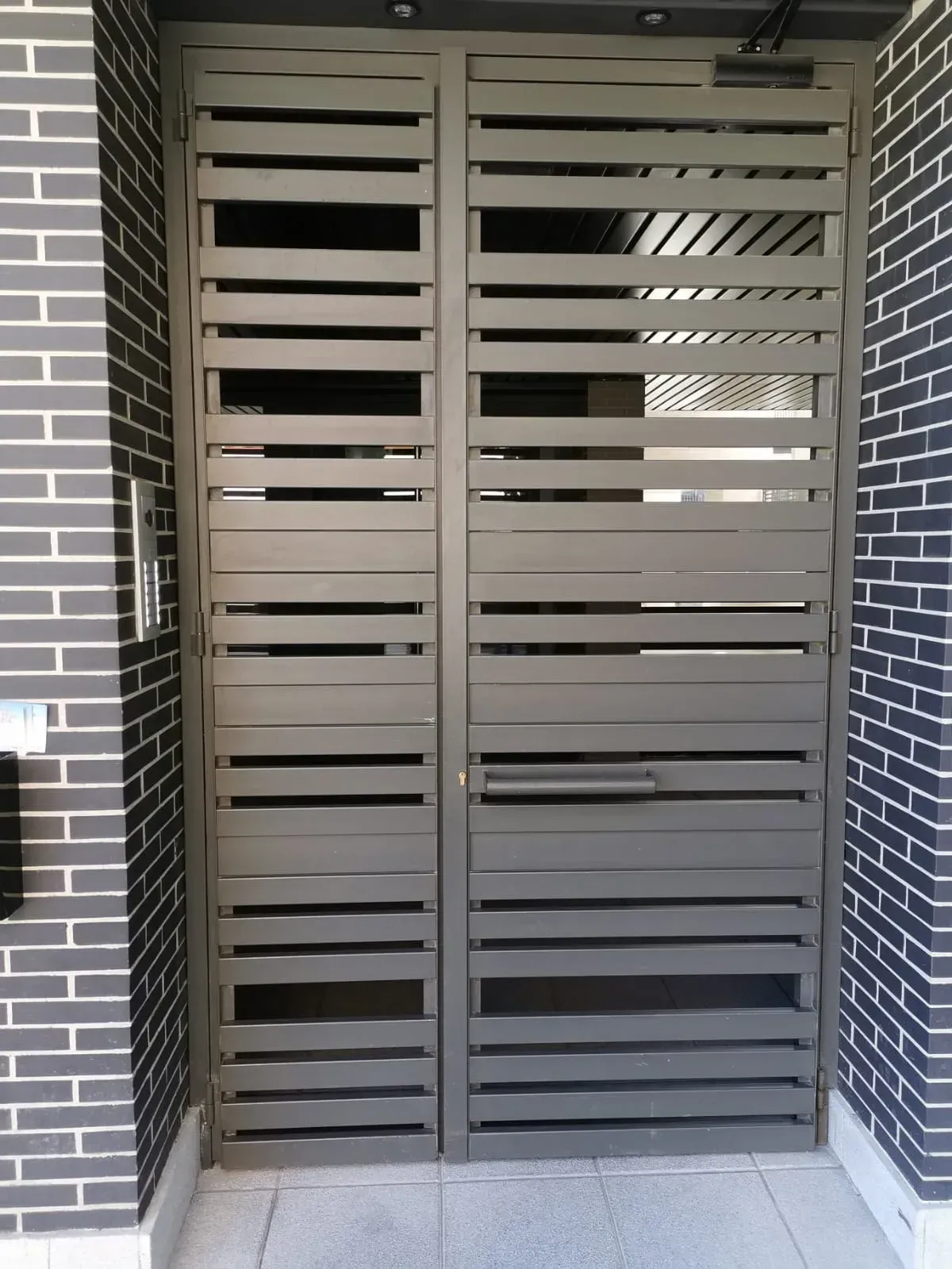 Puerta de lamas metálicas con rejillas de ventilación horizontales en una entrada revestida de ladrillos.