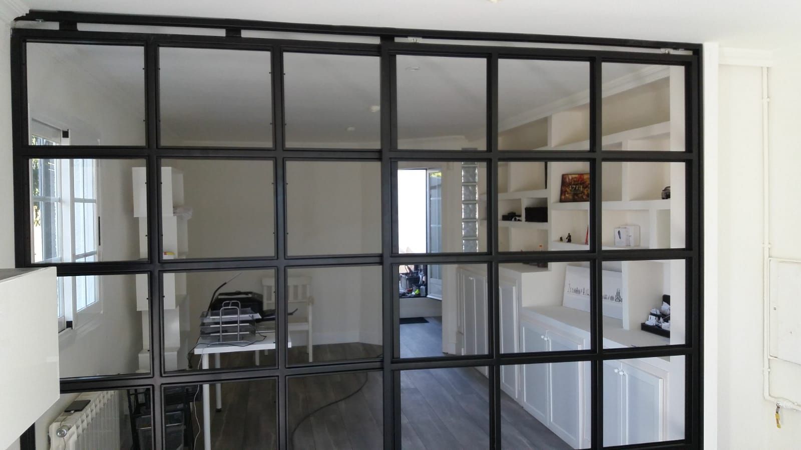 Una mampara de cristal con marco negro deja ver una habitación moderna con mesa, sillas y armarios blancos.
