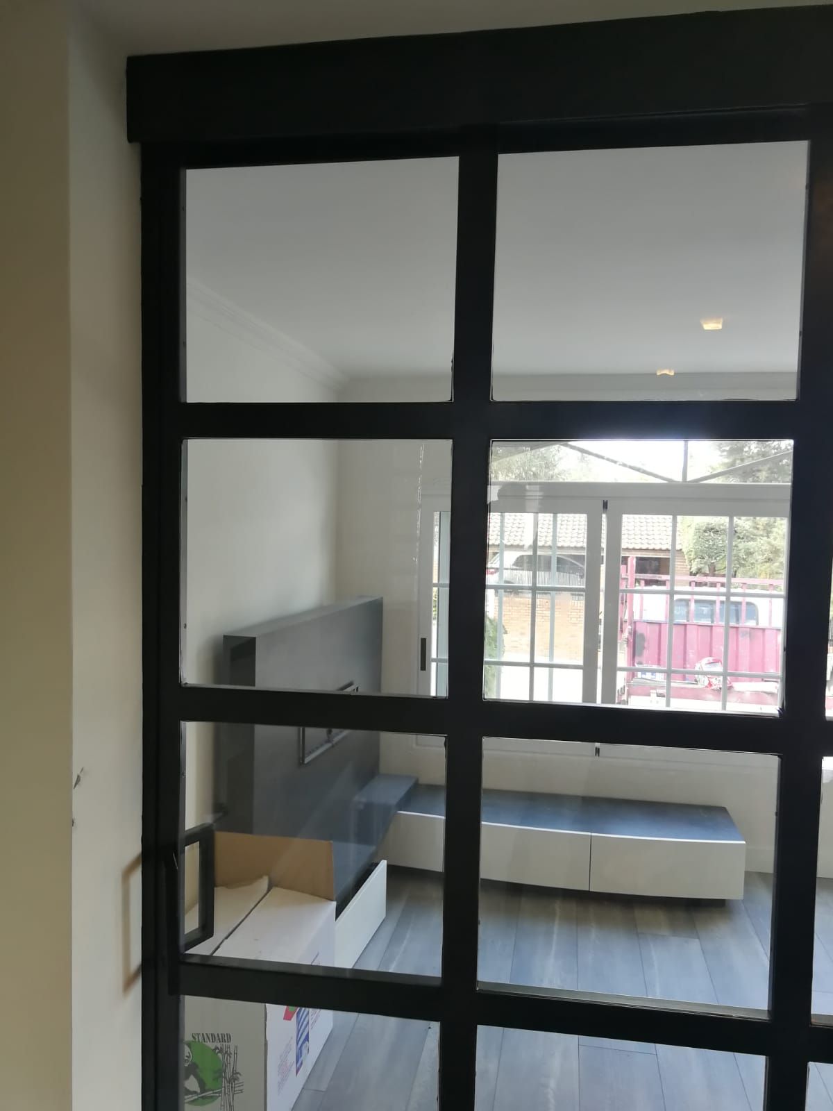 Ventana con marco negro que da a una habitación luminosa con televisor y asientos tipo banco.
