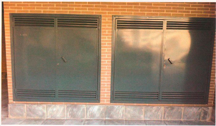 Dos contraventanas de metal oscuro incrustadas en una pared de ladrillo sobre una base de piedra.