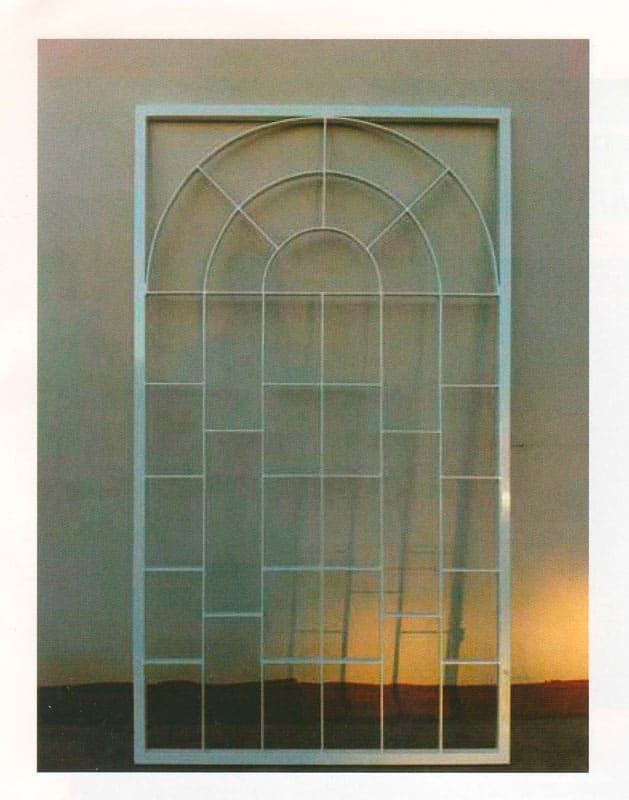 Reja decorativa de metal arqueada para ventana contra una pared beige con luz cálida en la parte inferior derecha.