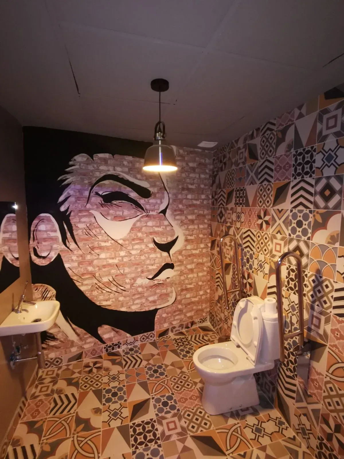 Baño pequeño con paredes de mosaico con estampados en negro, blanco y beige, y un gran mural con un rostro sobre el inodoro.
