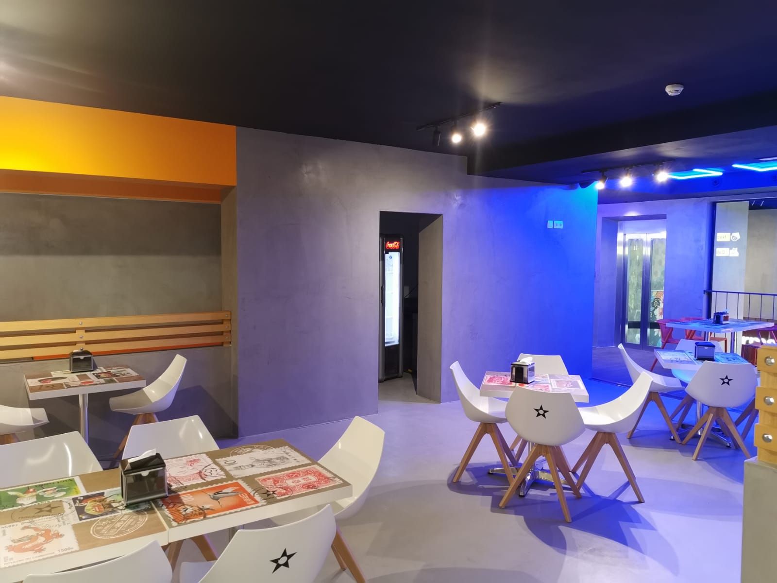 Interior de cafetería moderna con mesas y sillas blancas, iluminación cálida en tonos naranja y azul