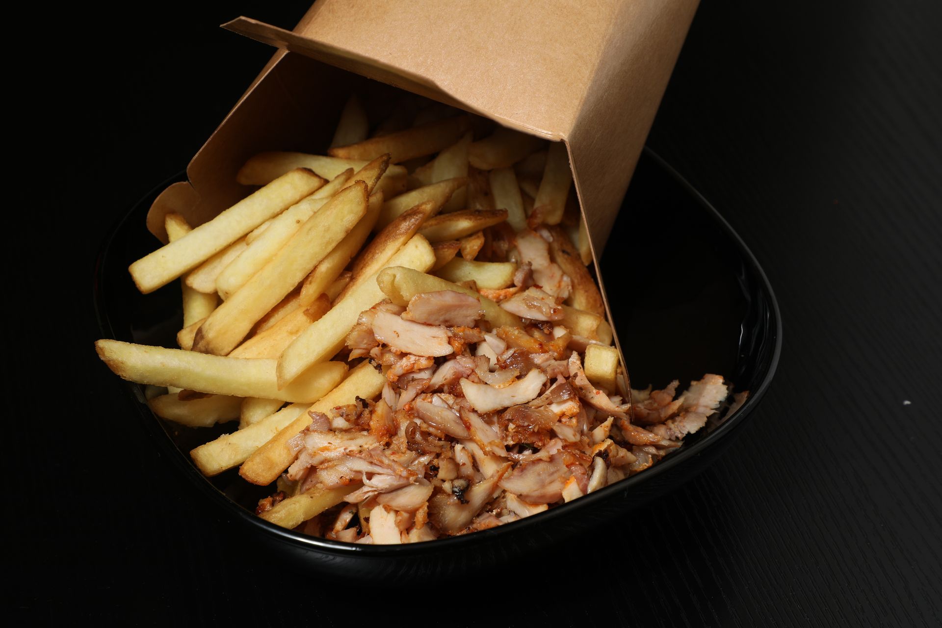 Une boîte en carton remplie de frites et garnie de viande effilochée, posée sur une assiette noire sur fond noir.