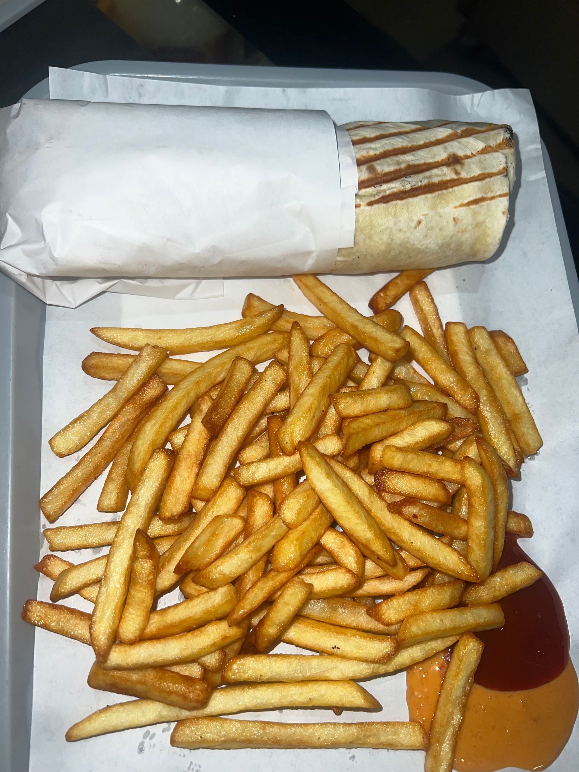 Un wrap grillé enveloppé dans du papier blanc est posé à côté d'un gros tas de frites dorées avec deux petites flaques de sauce.