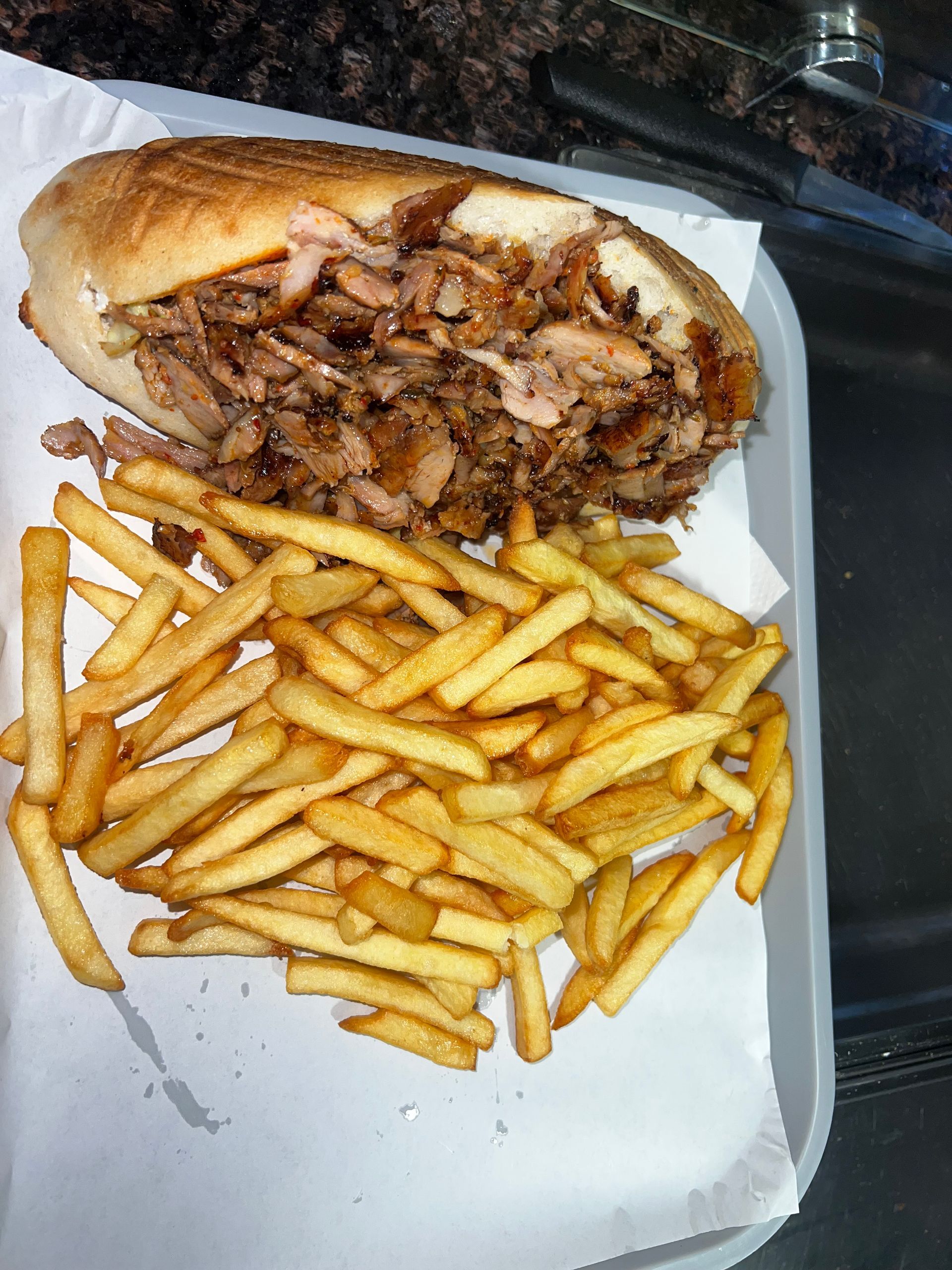 Un sandwich doner kebab garni, servi dans un petit pain avec une portion de frites dorées sur un plateau recouvert de papier blanc.