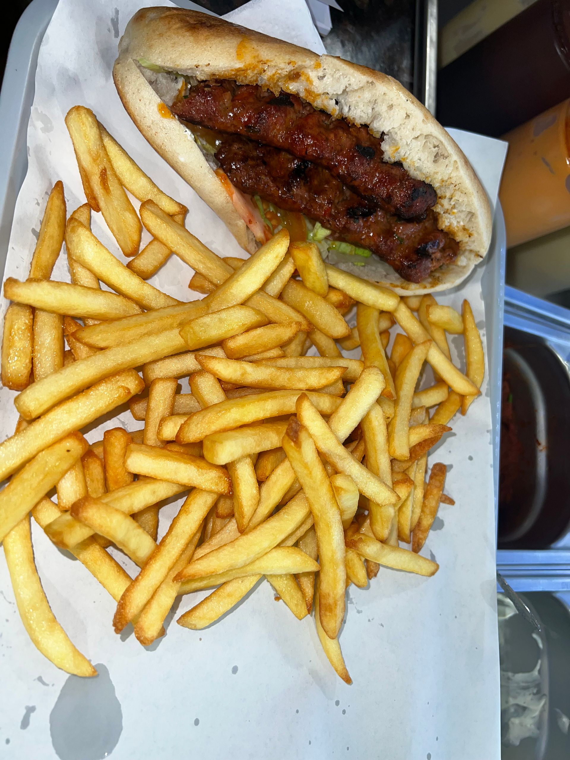 Un sandwich à la saucisse grillée sur un petit pain croustillant, servi avec une grande portion de frites dorées sur du papier sulfurisé.