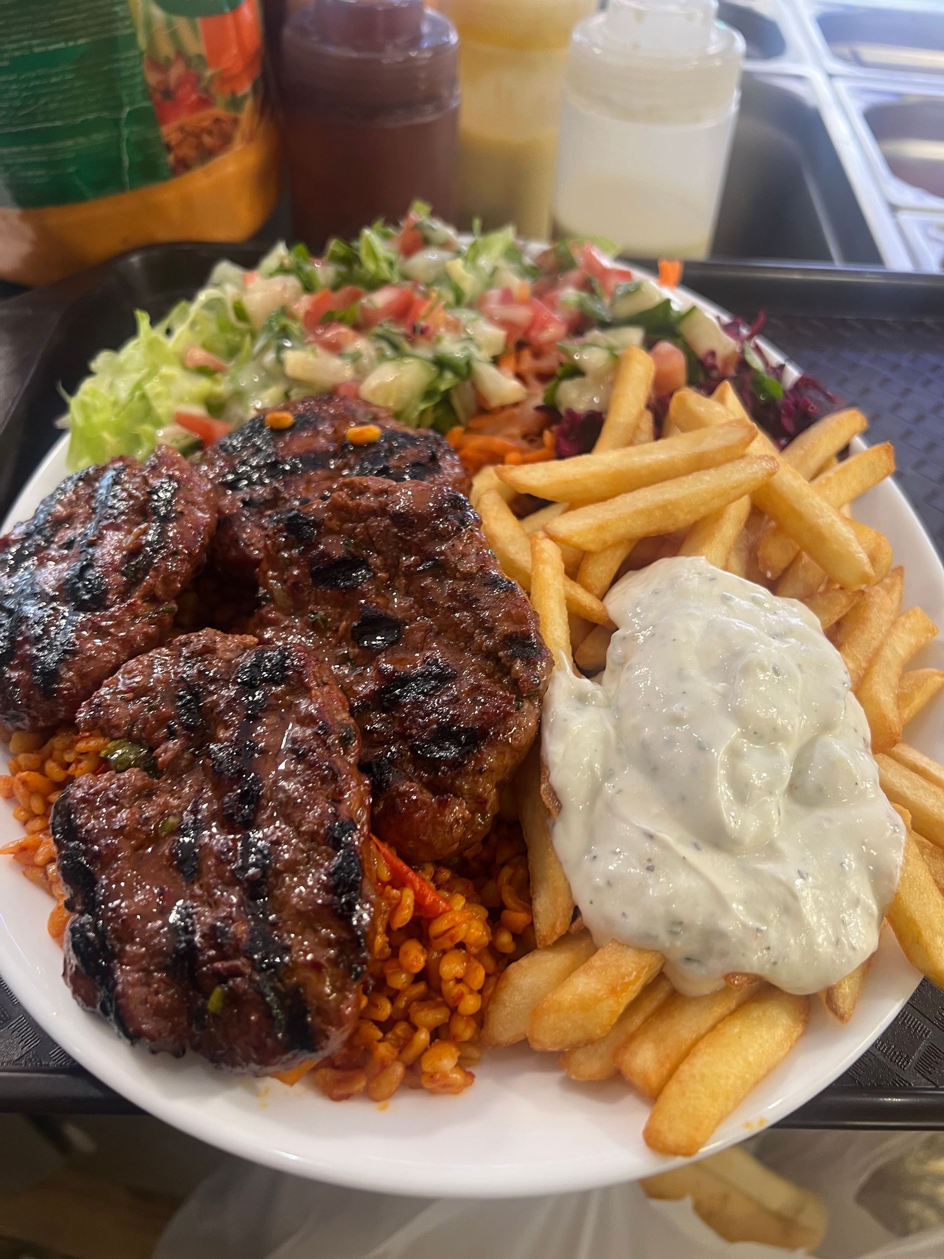 Une assiette de galettes de viande grillées servie avec des frites, du blé pilaf, une salade fraîche et une cuillerée de sauce tzatziki.