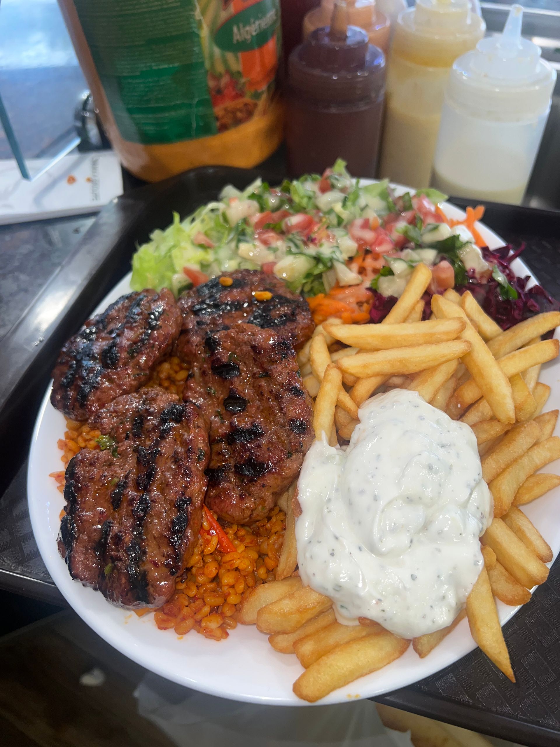 Une assiette de galettes de viande grillées, de blé, de frites et de salade fraîche, le tout nappé d'une généreuse cuillerée de sauce blanche.