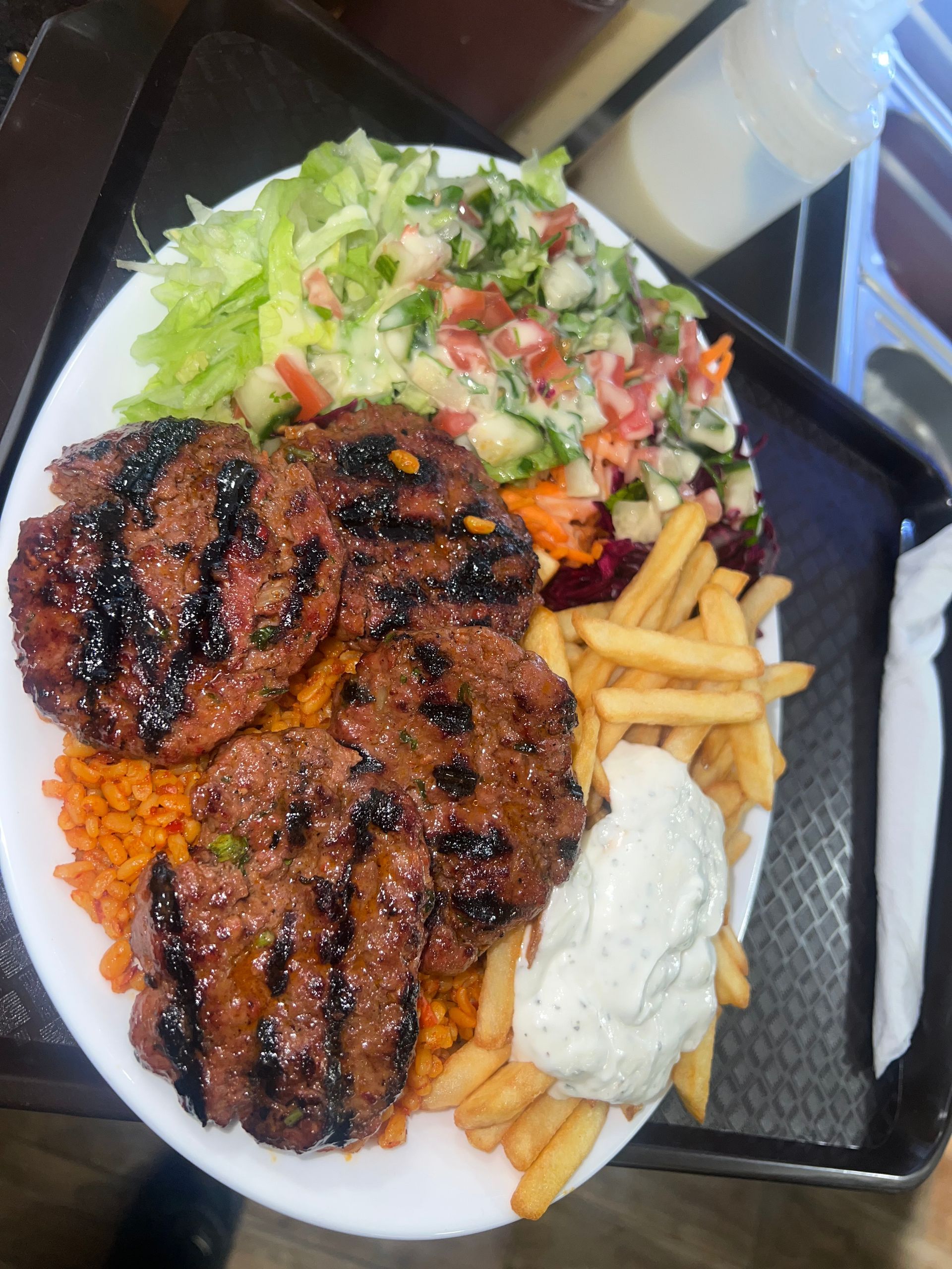 Une assiette de steaks hachés grillés servie avec des frites, du riz orange, une salade d'accompagnement et une cuillerée de sauce blanche.