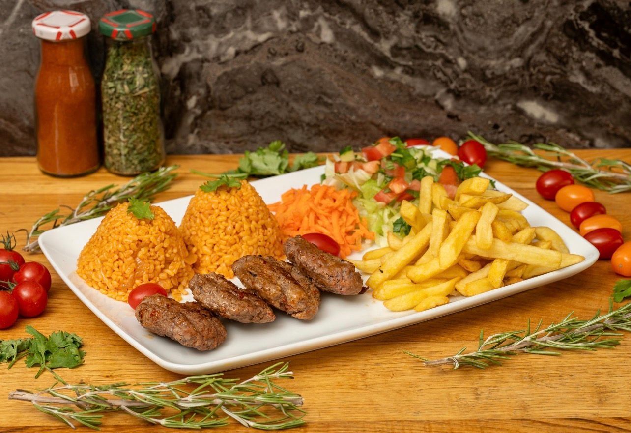Une assiette rectangulaire blanche garnie de boulettes de viande grillées, de deux monticules de riz orange, de carottes râpées, de salade et de frites.