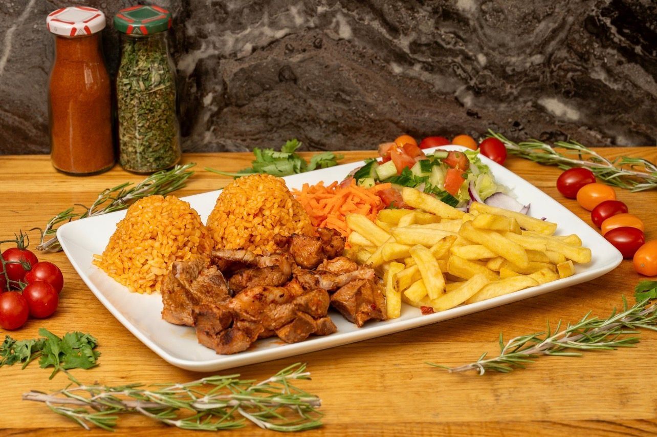 Sur une table rustique en bois, un plat blanc garni de morceaux de poulet, de riz jaune, de frites, de carottes râpées et d'une salade.