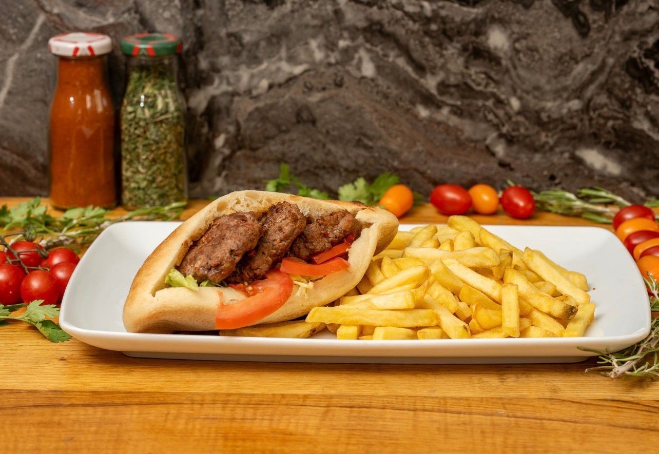 Un sandwich pita garni de viande grillée et de tranches de tomate, servi avec des frites sur une assiette rectangulaire blanche.