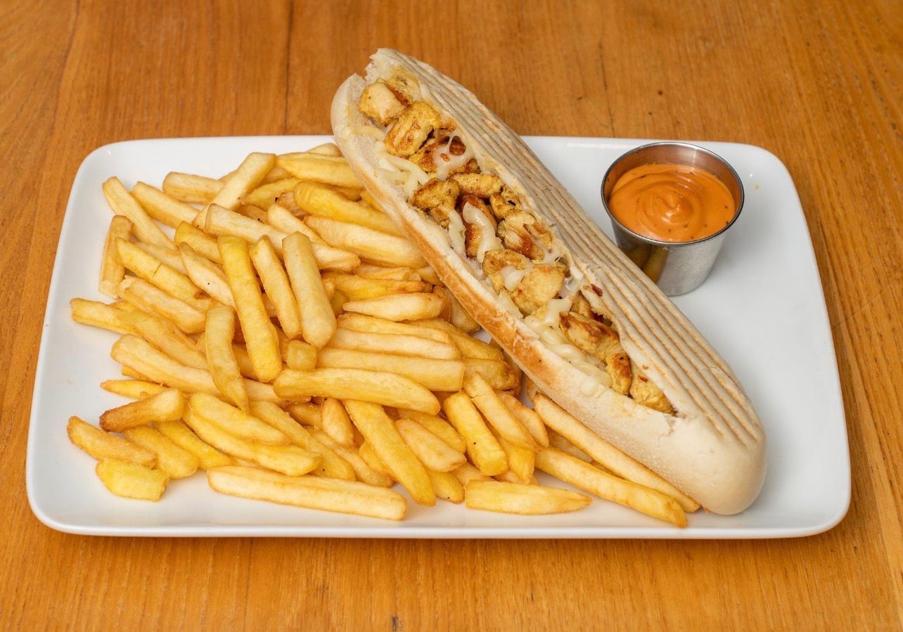 Une assiette rectangulaire blanche contenant un long panini au poulet, une grande portion de frites dorées et une petite coupelle de sauce.