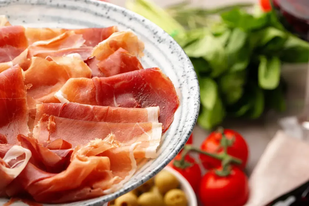 Primer plano de lonchas finas de jamón serrano sobre un plato texturizado, con tomates cherry y verduras al fondo.