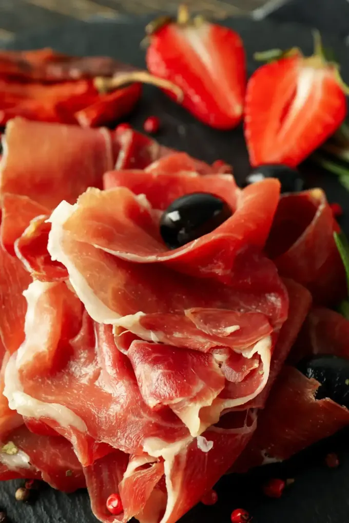 Lonchas de jamón serrano dispuestas sobre una superficie oscura con fresas partidas por la mitad.