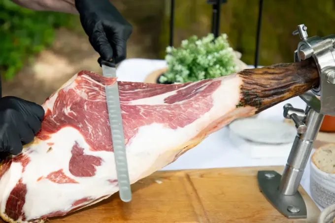 Una persona con guantes negros corta una pata de jamón curado que está sostenida.