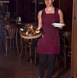 Un camarero con un delantal granate está de pie en un restaurante, sosteniendo una bandeja de aperitivos y una pila de platos pequeños.