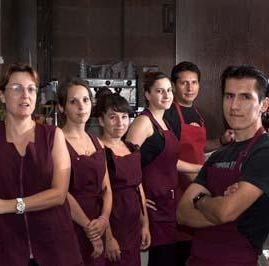Un grupo de cinco empleados, vestidos con delantales color burdeos y uniformes negros a juego, están de pie uno al lado del otro en la cocina de una cafetería.