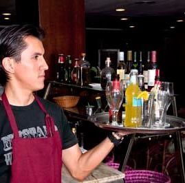 En un bar, un camarero con camisa negra y delantal rojo sostiene una bandeja plateada con vasos y botellas.