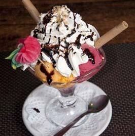 Un vaso para helado lleno de bolas de helado, cubierto con crema batida, sirope de chocolate, barquillos y una flor.