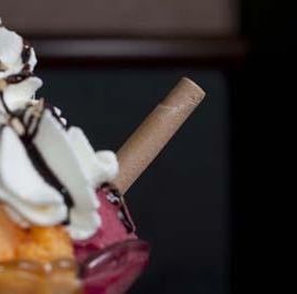 Primer plano de un helado con sabor a frutas, cubierto con crema batida, sirope de chocolate y una galleta de barquillo enrollada.