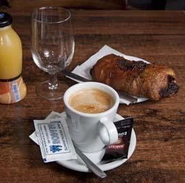 Un desayuno típico con una taza de café, un pastel de chocolate, un vaso vacío y una botella pequeña de zumo de naranja.