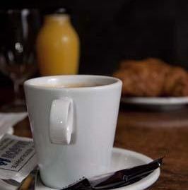 Una taza de café blanca reposa sobre un platillo, con cubiertos, un vaso de zumo y un cruasán al fondo.