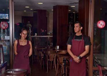 Dos empleados con delantales color granate están de pie en la entrada de un restaurante con decoración de madera.
