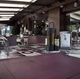 Una mesa en un comedor al aire libre, puesta con manteles individuales, aceiteras y un servilletero bajo un toldo de cristal.