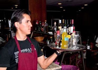 Un camarero con un delantal granate sostiene una bandeja con vasos y una botella de una bebida amarilla en un ambiente tenue, propio de un restaurante.