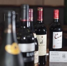 Una hilera de botellas de vino reposa sobre una superficie, con una pequeña carta en primer plano, sobre un fondo oscuro de interior.